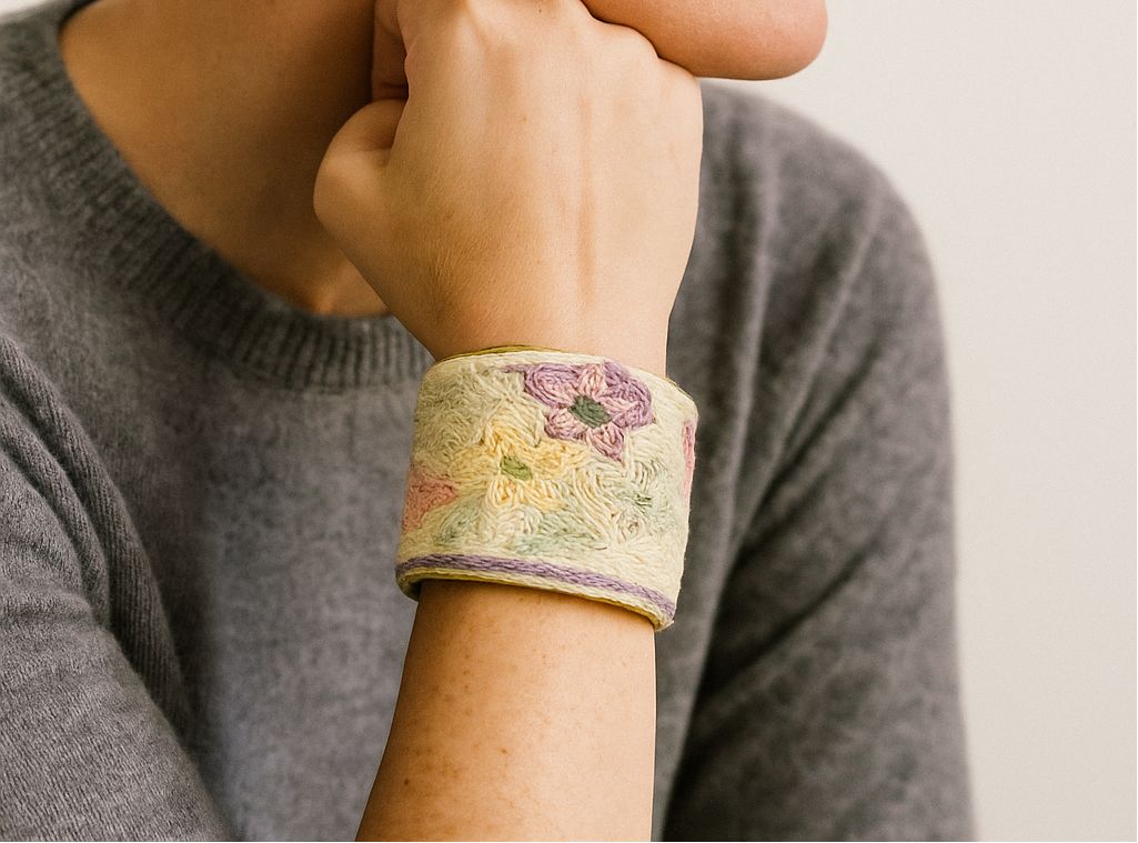 A female model wearing a one of a kind jewelry brass cuff bracelet for women—an open-back design wrapped in cream fabric with yellow and purple floral embroidery, showcased on her wrist against a neutral backdrop.