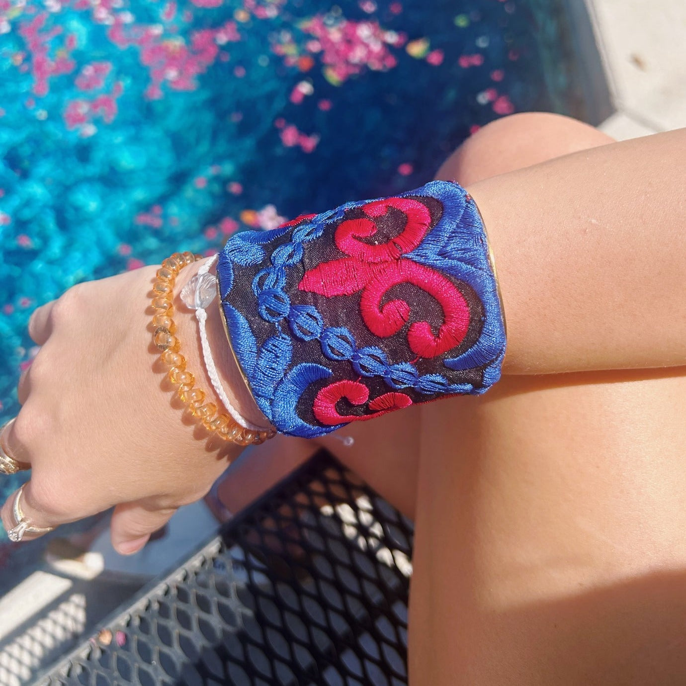 A female model wears a one of a kind jewelry brass cuff bracelet for women—a 3-inch wide, open-back design wrapped in black fabric with red and cobalt blue fleur-de-lis and scroll embroidery, shown on her wrist with a poolside background.