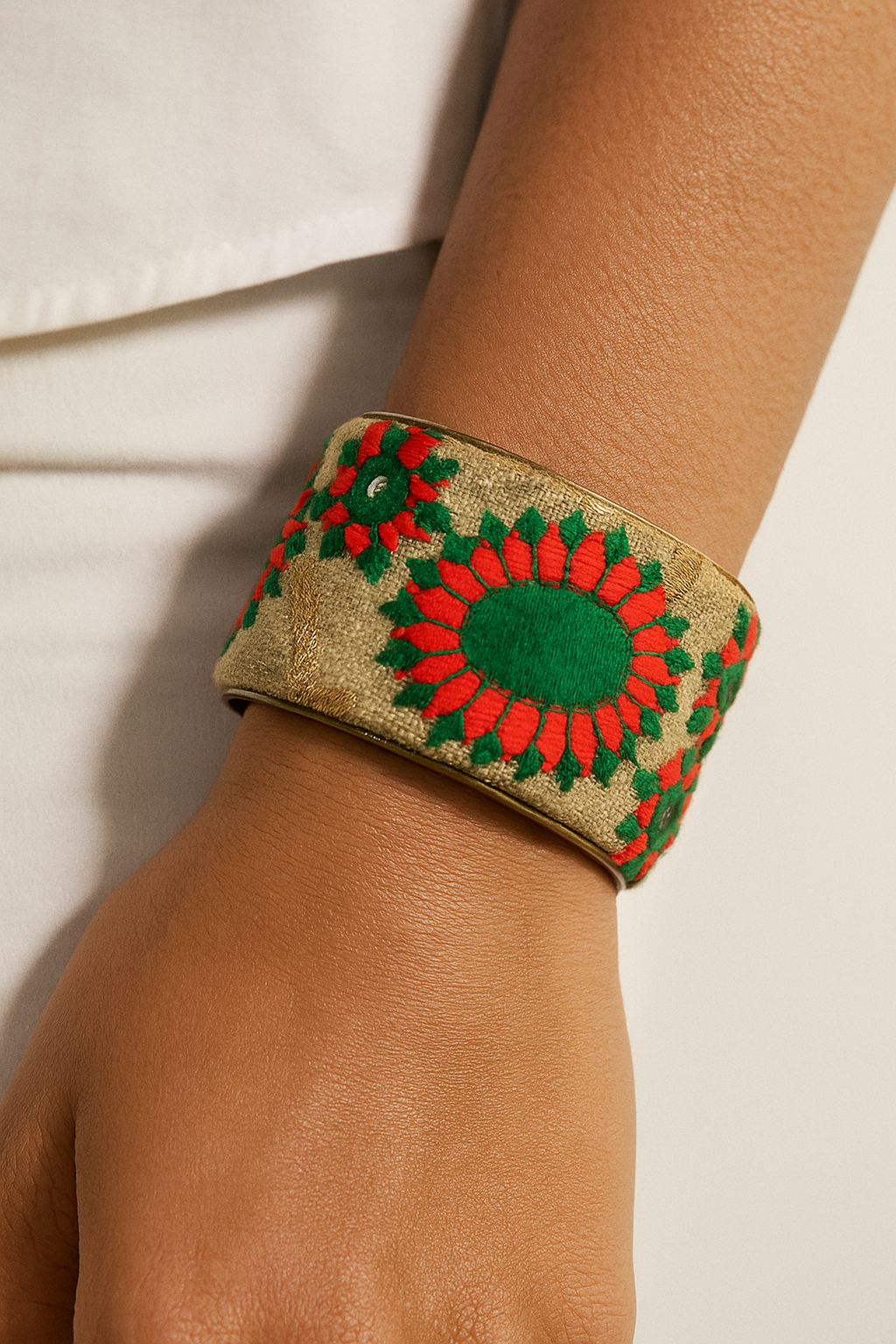 Close-up of a female model’s forearm and wrist wearing a 2-inch wide cream jacquard brass cuff bracelet embroidered with red floral motifs and green leaf accents; the open-back ends of the cuff are visible against a light background.