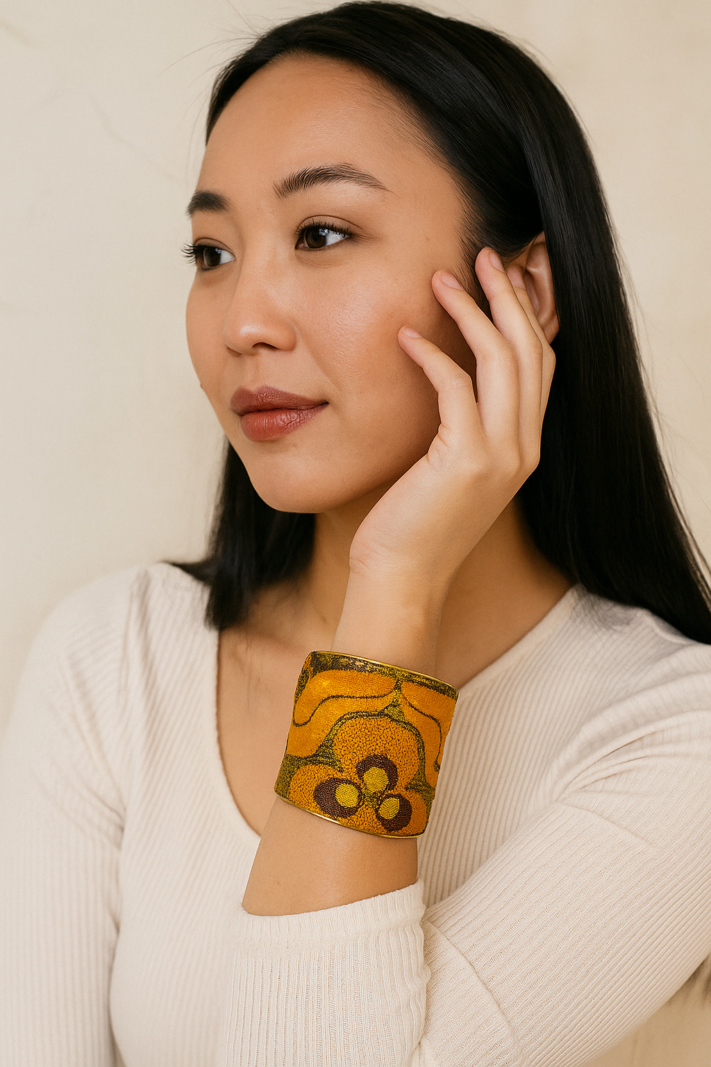 Close-up portrait of a female model wearing a 3-inch wide orange and gold fabric-wrapped brass cuff bracelet; the open-back ends peek out as she gently touches her face.
