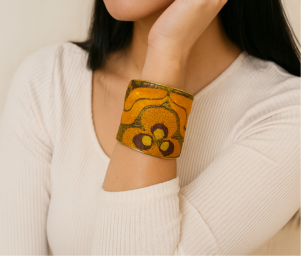 Close-up portrait of a female model wearing a 3-inch wide orange and gold fabric-wrapped brass cuff bracelet; the open-back ends peek out as she gently touches her face.