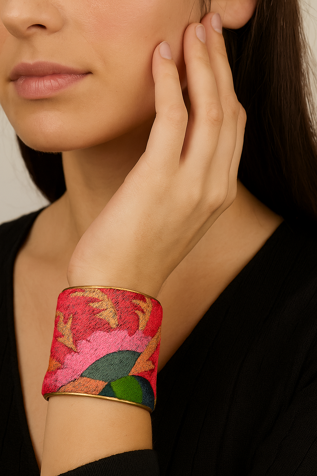 Close-up of a female model’s forearm and wrist wearing a 3-inch wide fuchsia and multicolor embroidered brass cuff bracelet; the open-back ends peek out as her hand rests near her face.