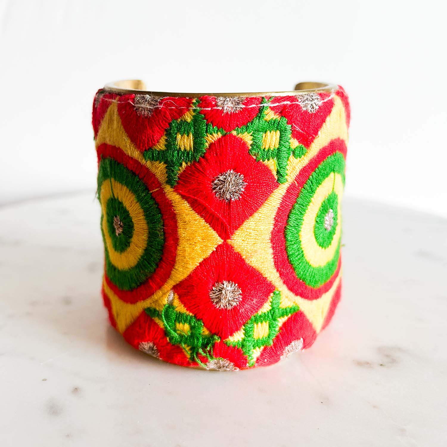 A standalone 3-inch wide raw brass cuff bracelet wrapped in plush velvet, featuring hand-embroidered concentric circle and diamond shapes in cherry red, lime green, sunshine yellow, and metallic silver threads on a white background.