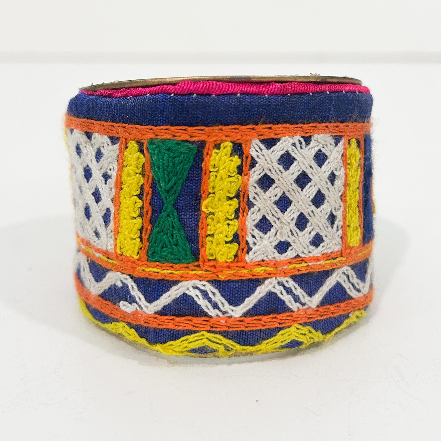 A standalone 2-inch wide raw brass cuff bracelet wrapped in deep cobalt-blue fabric embroidered with white lattice patterns, bordered by bright orange stitching, featuring vertical green hourglass motifs and yellow panels, finished with a slim pink trim along the top edge on a white background.