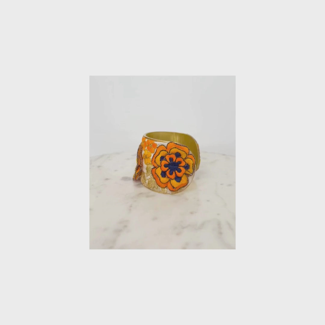 A gold flower cuff bracelet with orange and blue floral designs is displayed on a light marble surface against a white background.