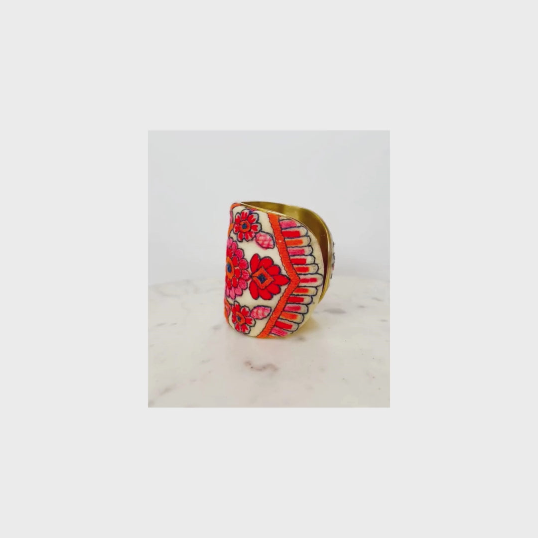 An embroidered cuff bracelet with red and pink floral designs on a pale yellow background is displayed elegantly on a marble surface.