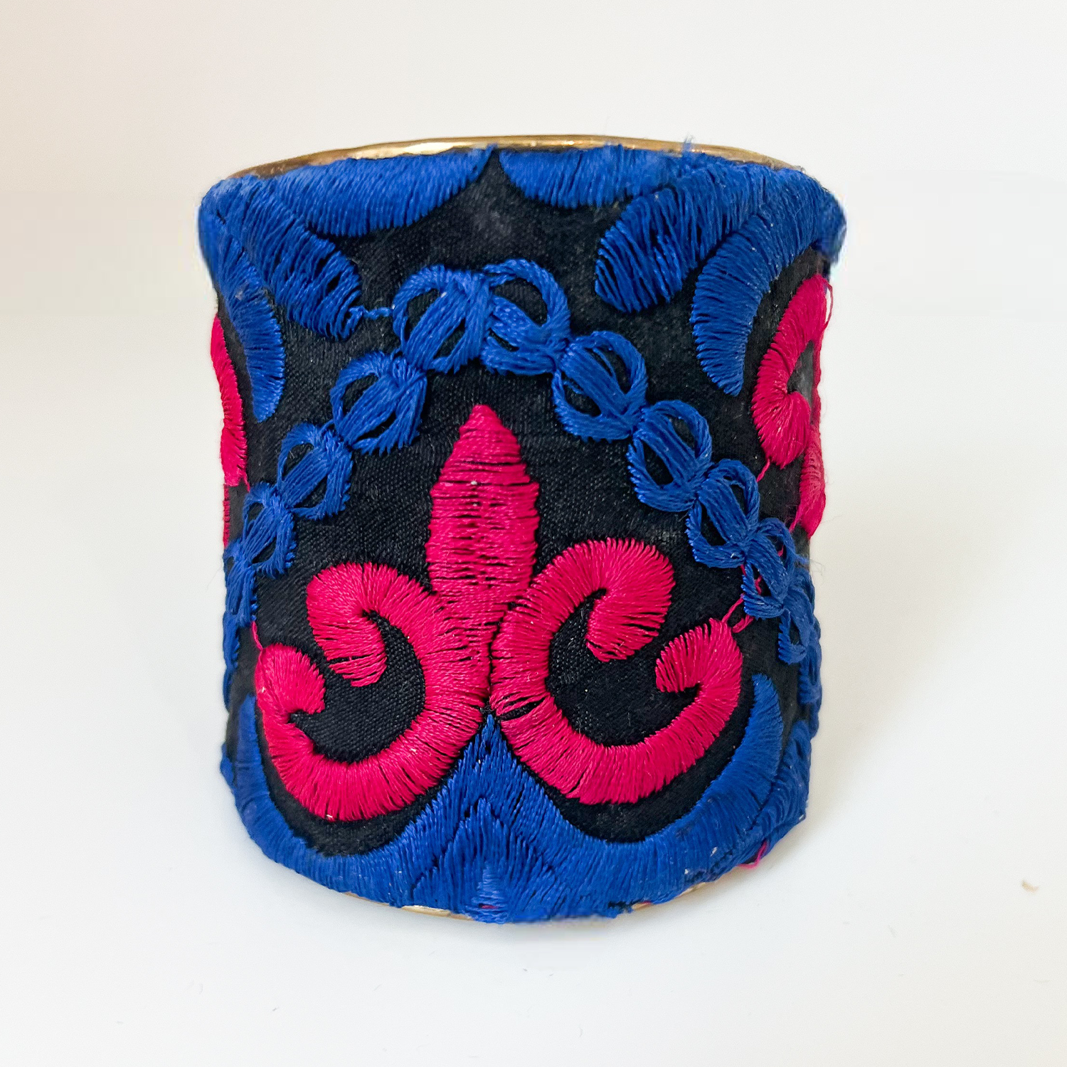 A studio image of a one of a kind jewelry brass cuff bracelet for women—a 3-inch wide, open-back raw brass cuff wrapped in black fabric with hand-embroidered red and cobalt blue fleur-de-lis and ornate scroll designs, displayed upright on a white surface.
