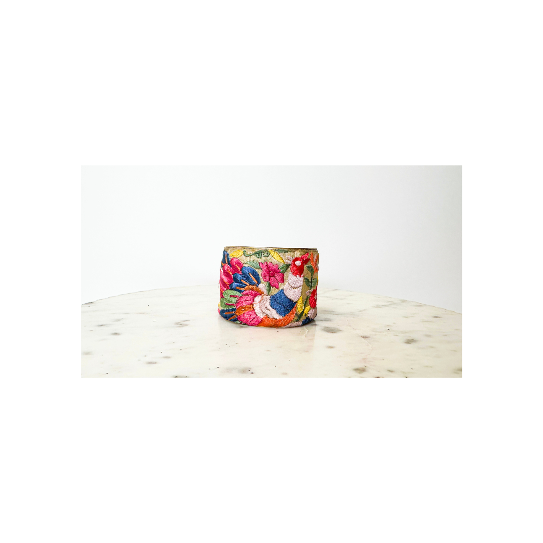 A studio image of a one of a kind jewelry brass cuff bracelet for women—a 2-inch wide, open-back design wrapped in navy fabric with multi-hued floral embroidery in pink, tangerine, emerald, and sapphire, displayed on a white marble surface.