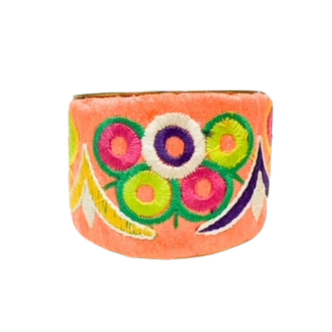A 2-inch wide raw brass cuff bracelet wrapped in coral velvet fabric, featuring hand-embroidered circular floral motifs with green, pink, yellow, and purple threads and stylized petal shapes on a white background.