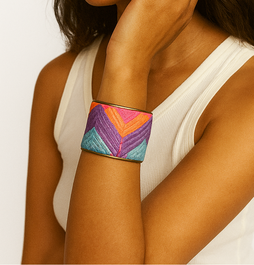 A woman wearing a 2-inch wide brass cuff bracelet wrapped in vibrant woven fabric featuring coral, teal, purple, and pink chevron stripes. Her arm is lifted near her ear, showing the cuff’s shimmer and fit.
