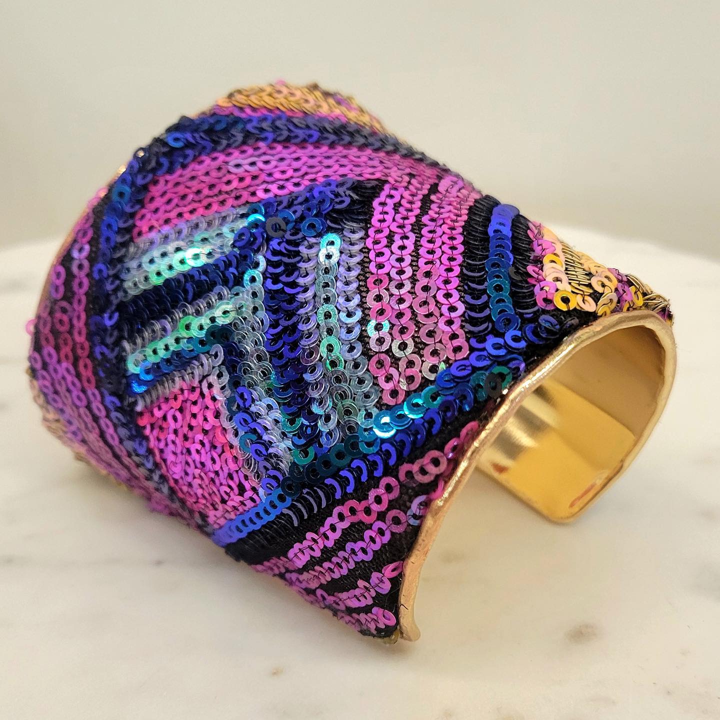A 3-inch wide raw brass cuff bracelet entirely covered in iridescent sequins—fuchsia, violet, teal, black, and gold—arranged in diagonal stripe patterns, photographed at an angle to show the open-back and gleaming brass interior on a white marble surface.